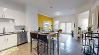 This thoughtfully designed 1-bedroom, 1-bathroom unit offers an efficient and stylish layout across a single floor. The open-concept kitchen and living area serves as the heart of the space, featuring dark hardwood-style flooring, crown molding, and abundant natural light from large windows and French doors. The kitchen is equipped with stainless steel appliances — including a refrigerator, microwave, and dishwasher — complemented by crisp white cabinetry, a tile backsplash, and a built-in dining bar with seating. A bold golden-yellow accent wall anchors the living area, adding a vibrant focal point and visual warmth to the otherwise neutral palette. The bedroom is a serene retreat with windows that invite natural light, continuing the dark hardwood flooring, and direct access to a walk-in closet outfitted with built-in shelving and hanging rods. The full bathroom features a dark granite vanity countertop, white cabinetry, and a soaking tub with shower. Additional conveniences include a dedicated laundry area and pantry storage. Crown molding throughout and track lighting in the kitchen add refined architectural detail to the overall design.
