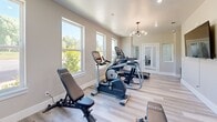 Located at 651 Osigian Blvd in Warner Robins, Georgia, this dedicated exercise room is a fully appointed fitness space designed to support a comprehensive workout routine. The room features light wood-tone flooring throughout, lending a clean and modern aesthetic that complements the neutral, warm-gray wall tones and bright white trim and baseboards.

Natural light flows generously into the space through multiple large windows, offering views of the surrounding greenery and creating an inviting, energizing atmosphere. A stylish globe-cluster chandelier serves as a central design focal point, complemented by recessed lighting to ensure consistent illumination throughout the room.

The exercise room is outfitted with an impressive array of commercial-grade fitness equipment, including Matrix-brand cardio machines such as a treadmill, elliptical, and stationary bike, as well as a multi-function cable strength training machine. A full dumbbell rack with a comprehensive weight selection rounds out the strength training offerings, while adjustable workout benches provide versatility for a variety of exercises.

A wall-mounted flat-screen television adds a functional entertainment element, and a set of glass-paneled French doors connects the space to an adjacent area, enhancing the open and airy feel. The layout is thoughtfully organized to maximize both equipment accessibility and ease of movement throughout the room.