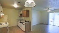 This single-story space presents an open-concept layout where the kitchen flows directly into an adjacent living area, creating a connected and functional floor plan. The kitchen is equipped with a continuous countertop that includes an integrated sink and a built-in dishwasher. Upper cabinetry in warm wood tones is complemented by white lower cabinets, providing a contrast that adds visual interest to the space. A prominent dome pendant light fixture hangs above the kitchen area, serving as a focal point and providing direct overhead illumination. A secondary flush-mount ceiling light further brightens the space. The floors throughout are covered in light-toned tile, offering a cohesive look between the kitchen and the open living area. The walls are finished in a neutral off-white, contributing to a clean and understated aesthetic. The textured ceiling is consistent across both areas. The living area, visible beyond the kitchen, is an open, flexible space well-suited for various furniture configurations. Electrical outlets are positioned along the walls, and HVAC vents are integrated into both the ceiling and walls for climate control.