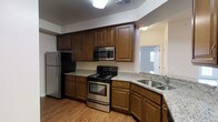 This single-floor space is designed with practicality and comfort in mind. The kitchen serves as a central highlight, showcasing rich, warm-toned wooden cabinetry that extends from floor to ceiling, providing generous storage throughout. The countertops feature a speckled granite-style surface that complements the cabinetry beautifully. Stainless steel appliances, including a refrigerator, electric range, and over-the-range microwave, lend a modern, cohesive look to the space. The double-basin stainless steel sink is positioned at the corner of the counter, offering a practical and ergonomic workspace. Light-toned hardwood-style flooring runs throughout, adding warmth and visual continuity. The kitchen layout follows a functional L-shape design, maximizing counter and storage space while maintaining an open, easy-to-navigate flow. Overhead lighting ensures the workspace is well-lit, and a passthrough view into an adjacent room hints at a connected, open layout. The overall ambiance is clean, practical, and inviting, making this space well-suited for everyday living.