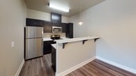 This single-floor residence offers a well-organized layout with distinct living areas, bedrooms, and bathrooms. The kitchen serves as a functional centerpiece, outfitted with dark espresso cabinetry, white countertops, and stainless steel appliances including a refrigerator, electric range, and microwave. A peninsula with bracket-supported overhang provides casual seating and separates the kitchen from the adjoining area. Wood-look flooring and a pendant light add a contemporary touch. The living area features plush carpeting, a built-in wall shelf for display or storage, and sliding glass doors that invite natural light and outdoor access. Multiple carpeted bedrooms offer a calm, neutral palette with taupe walls, white trim, and windows fitted with horizontal blinds that frame views of surrounding greenery. One bedroom includes a ceiling fan and a closet. The bathrooms provide two distinct bathing options: one features a full tub and shower combination with white tile surround, while the other includes a walk-in shower stall with chrome fixtures and an adjacent dark vanity with a light countertop. Wood-look tile flooring completes the bathroom spaces, maintaining a cohesive aesthetic throughout the residence.