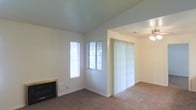 This single-floor space presents a clean, functional layout centered around a carpeted living area with neutral white walls and vaulted ceilings that lend a sense of openness and height. The living room is anchored by a fireplace along one wall, adding a practical and inviting focal point to the space. Two windows, including a tall narrow window and a standard horizontal window fitted with blinds, allow natural light to filter into the room, brightening the interior throughout the day.

An open archway connects the living area to an adjacent room, which features vertical blinds covering what appears to be a sliding glass door — likely providing access to an outdoor area. A ceiling fan with a light fixture is visible in this connected room, contributing to airflow and additional lighting. The consistent use of neutral tones throughout — from the white walls to the beige carpet — creates a cohesive, understated aesthetic. The open-plan connection between rooms enhances the sense of flow and usability, making the space feel well-integrated and practical for everyday living.