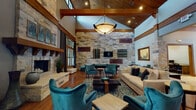 This beautifully designed single-floor space makes an immediate impression with its dramatic vaulted ceilings and rich exposed wood beam framework. The main living area centers around a floor-to-ceiling natural stone fireplace, complete with a substantial wood mantel and raised stone hearth, serving as a striking architectural focal point. Abundant natural light pours in through multiple wood-framed windows adorned with floor-length drapes, offering views of the surrounding greenery. Warm hardwood flooring flows throughout, complementing the earthy stone accents and rich wood trim that define the interior's character. The open layout connects the living area seamlessly to a game room featuring a pool table and a full stone accent wall with a mounted television. A well-appointed kitchen area includes stainless steel appliances, warm wood cabinetry with glass fronts, mosaic tile backsplash, a generous white countertop bar, and pendant lighting. The welcoming entry foyer showcases a stone column detail, cool blue-grey walls, and curated artwork. Throughout the space, teal velvet seating, layered area rugs, and carefully selected décor create a cohesive, upscale yet inviting ambiance that balances rustic craftsmanship with modern comfort.