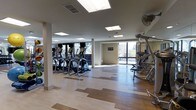 This modern fitness center is designed to provide a comprehensive workout environment within a single open-plan floor. The space is anchored by a row of elliptical cardio machines lined along one side, complemented by a multi-station strength training apparatus on the opposite end, offering users a full range of exercise options. The flooring features a distinctive mixed-tone hardwood pattern, combining light and dark wood tones in a geometric arrangement that adds visual interest while remaining practical for athletic use. Natural light floods the interior through expansive floor-to-ceiling windows along the perimeter, offering views of the surrounding outdoor landscape and creating a bright, energizing atmosphere. Overhead, a series of recessed skylights and flush-mounted ceiling fixtures provide consistent, even illumination throughout the space. The ceiling is clean and contemporary, with visible ventilation grilles ensuring proper air circulation. Neutral-toned walls contribute to a calm, focused environment, while a ceiling fan near the strength equipment area adds additional airflow. The overall layout is open and uncluttered, allowing for easy movement between equipment zones and providing ample floor space for stretching or additional exercises.