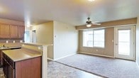 This single-floor space offers a thoughtfully designed open-concept layout that seamlessly connects the kitchen and living areas. The living room is generously sized, featuring plush wall-to-wall carpeting that adds warmth and comfort underfoot. A ceiling fan with a built-in light fixture is centrally positioned to provide both airflow and ambient lighting throughout the space. A large picture window floods the room with natural light and frames views of the surrounding exterior, while also providing access to a private balcony — ideal for enjoying the outdoors. The kitchen area transitions smoothly from the living space, marked by a raised bar-height counter with rich dark wood cabinetry, offering a casual seating or prep area. Hard-surface tile flooring defines the kitchen zone, creating a practical and easy-to-maintain surface. Neutral-toned walls throughout lend a clean and versatile backdrop, making the space adaptable to a variety of interior styles. A hallway visible from the main area suggests additional rooms beyond, contributing to the overall functional flow of the floor plan.