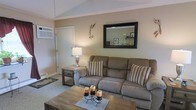 This single-floor home offers a comfortable and well-appointed living environment with a welcoming ambiance throughout. The living room serves as the heart of the space, featuring vaulted ceilings that lend an airy, open feel to the room. Neutral warm beige walls create a calm and versatile backdrop, complemented by plush carpeting underfoot. A generously sized reclining sofa anchors the seating area, styled with striped accent pillows that add a subtle touch of pattern and color. A rustic wooden coffee table sits at the center, accented with candle décor, while a matching end table and a classic table lamp provide soft, ambient lighting. A large framed wall mirror above the sofa reflects light and enhances the sense of depth in the room. Decorative wall accents and a baseboard heating unit are also present, adding to the room's functional character. A white interior door provides access to additional areas of the home. The overall design aesthetic is cozy and understated, blending practical furnishings with warm, homey décor to create a space that feels lived-in and inviting.
