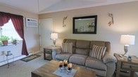 This single-floor home offers a comfortable and well-appointed living environment with a welcoming ambiance throughout. The living room serves as the heart of the space, featuring vaulted ceilings that lend an airy, open feel to the room. Neutral warm beige walls create a calm and versatile backdrop, complemented by plush carpeting underfoot. A generously sized reclining sofa anchors the seating area, styled with striped accent pillows that add a subtle touch of pattern and color. A rustic wooden coffee table sits at the center, accented with candle décor, while a matching end table and a classic table lamp provide soft, ambient lighting. A large framed wall mirror above the sofa reflects light and enhances the sense of depth in the room. Decorative wall accents and a baseboard heating unit are also present, adding to the room's functional character. A white interior door provides access to additional areas of the home. The overall design aesthetic is cozy and understated, blending practical furnishings with warm, homey décor to create a space that feels lived-in and inviting.