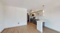 Located at 10720 5th Avenue NE in Seattle's Northgate neighborhood, this single-floor unit offers a clean, contemporary design throughout. The open-concept living and kitchen area is anchored by warm wood-tone luxury vinyl plank flooring and crisp white walls, creating a bright and welcoming atmosphere. Large sliding glass doors lead to a private balcony, flooding the living space with natural light and offering views of the surrounding Seattle skyline and neighborhood.

The kitchen is well-appointed with a two-tone cabinetry scheme — pairing dark espresso lower cabinets with light upper cabinets — complemented by white countertops and a breakfast bar peninsula ideal for casual dining. Stainless steel appliances include a refrigerator, electric range, microwave, and dishwasher. Track lighting and a pendant light fixture add both function and style overhead.

The bathroom features textured tile flooring with an earthy, slate-like appearance, a full tub/shower combination with a grab bar, a wood-toned vanity, and white countertops. A dedicated in-unit laundry area houses a stacked full-size washer and dryer, providing excellent everyday convenience. The overall design is modern, clean, and move-in ready.
