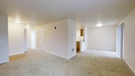This single-floor residential unit offers a clean, functional layout designed for comfortable everyday living. The main living area is generously proportioned, featuring soft, neutral-toned wall-to-wall carpeting that adds warmth underfoot. Crisp white walls and white trim throughout the space create a bright, airy atmosphere, complemented by flush ceiling lighting that ensures even illumination across the room.

A central dividing wall partially separates the living area from the kitchen, creating a sense of definition between the two spaces while maintaining an open, connected feel. The kitchen features warm wood-toned cabinetry, a countertop with a built-in sink, and hard flooring that contrasts neatly with the carpeted living spaces. A ventilation register is positioned at the base of the dividing wall, indicating a built-in climate control system throughout the unit.

A paneled interior door is visible along one wall, providing access to an adjacent room or hallway, while a secondary doorway leads further into the unit. The overall design is straightforward and practical, with a neutral color palette of beige and white tones that offers a versatile backdrop suitable for a range of furnishing styles. The space presents a move-in-ready environment with a cohesive, understated aesthetic.