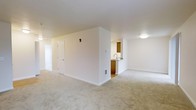 This single-floor residential unit offers a clean, functional layout designed for comfortable everyday living. The main living area is generously proportioned, featuring soft, neutral-toned wall-to-wall carpeting that adds warmth underfoot. Crisp white walls and white trim throughout the space create a bright, airy atmosphere, complemented by flush ceiling lighting that ensures even illumination across the room.

A central dividing wall partially separates the living area from the kitchen, creating a sense of definition between the two spaces while maintaining an open, connected feel. The kitchen features warm wood-toned cabinetry, a countertop with a built-in sink, and hard flooring that contrasts neatly with the carpeted living spaces. A ventilation register is positioned at the base of the dividing wall, indicating a built-in climate control system throughout the unit.

A paneled interior door is visible along one wall, providing access to an adjacent room or hallway, while a secondary doorway leads further into the unit. The overall design is straightforward and practical, with a neutral color palette of beige and white tones that offers a versatile backdrop suitable for a range of furnishing styles. The space presents a move-in-ready environment with a cohesive, understated aesthetic.