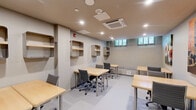 Located at 525 Main St in Hartford, Connecticut, this single-floor office space offers a efficiently organized and fully functional work environment. The room accommodates multiple light wood-topped workstations paired with ergonomic mesh-back rolling chairs, arranged to maximize the available floor area and support both individual and collaborative work. Recessed ceiling lighting throughout ensures consistent, even illumination across the entire space, while a centrally mounted cassette-style HVAC unit provides climate control year-round. Along the walls, open-faced wooden wall-mounted shelving units provide practical storage and display options at multiple heights. A low storage credenza with colorful locker-style sliding doors adds a touch of personality and additional storage near one wall. Natural light filters in through a row of ground-level windows along one wall, offering a subtle connection to the outdoors. The space is further enlivened by a curated selection of colorful wall art, including vibrant canvas prints depicting floral and urban motifs. Neutral-toned walls and a smooth light gray floor finish contribute to a clean, professional aesthetic. A padded bench seat beneath the windows provides an additional informal seating option. The overall ambiance is practical and organized, well-suited for a focused, productive work setting.