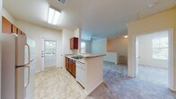 This thoughtfully designed residential unit offers a comfortable and functional layout across its levels, connected by a carpeted staircase with a clean white handrail. The open-concept kitchen and living area form the heart of the home, where warm reddish-brown cabinetry, granite-style countertops, and white appliances — including an electric range, built-in microwave, and dishwasher — create a practical and inviting cooking space. Light tile flooring runs throughout the kitchen, which flows seamlessly into a spacious living area finished with plush neutral carpeting, crown molding, and a ceiling fan. A peninsula counter separates the two spaces while maintaining an open, airy feel. Natural light pours in through multiple windows fitted with horizontal blinds, brightening each room. The bedroom features soft carpeting, a large window with an outdoor view, and ample wall space. The bathroom is finished with tile flooring, a vanity with a granite-style countertop, and clean white fixtures. A tiled utility or transition area with large windows adds additional versatility to the layout. Throughout, the warm cream-toned walls and consistent finishes create a cohesive, welcoming ambiance suited for comfortable everyday living.