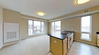 This single-floor space features an open-concept layout that seamlessly connects the kitchen and living areas. The kitchen is anchored by a peninsula with a dark countertop, an undermount double sink with a modern faucet, and light wood cabinetry below, providing ample storage. A dishwasher is integrated into the peninsula, enhancing everyday functionality. The transition from tile flooring in the kitchen area to wall-to-wall carpeting in the living space creates a clear visual distinction between zones while maintaining a cohesive feel.

The living area is generously proportioned, offering flexible space for furniture arrangement. Large windows along two walls flood the interior with natural light and frame views of the surrounding neighborhood and bare winter trees, giving the space an airy, open character. The neutral beige walls and white ceiling complement the flush-mount ceiling light, which adds a clean, understated touch. Subtle architectural detailing, including a crown molding-style border near the ceiling, adds a refined element to the otherwise straightforward design. Overall, the space presents a practical and well-lit environment with a calm, neutral aesthetic.