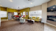 This single-floor community space offers a seamlessly integrated living room, dining area, and kitchen designed for comfort and functionality. The space is immediately welcoming, featuring a glass-paneled entry door that floods the foyer with natural light, complemented by tile flooring and a dark wood console station near the entrance.

The living area is anchored by a plush sofa, accent chairs, and a wall-mounted flat-screen TV alongside a sleek electric fireplace insert, all set against a warm taupe wall. Bold chartreuse green accent walls create a lively visual contrast throughout the open layout. Multiple windows with horizontal blinds bring in ample natural light and views of the surrounding outdoor landscape.

The dining area features a high-top communal table with bar-height seating beneath a contemporary geometric pendant light, creating an ideal space for group gatherings. The kitchen is equipped with stainless steel appliances — including a refrigerator, range, microwave, and dishwasher — paired with wood-grain cabinetry and dark marble-style countertops. A window above the sink enhances the kitchen's brightness.

Flooring transitions smoothly between tile in the kitchen and entry areas and textured carpet in the living and dining spaces. Recessed lighting throughout maintains a clean, modern ceiling line, completing this well-appointed communal setting.
