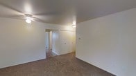 This single-floor space presents a straightforward and functional layout with a neutral, move-in-ready aesthetic. The interior is finished with wall-to-wall carpeting in a warm beige tone, providing a comfortable underfoot feel throughout the main living area. The walls are painted in a clean, bright white, lending an open and airy atmosphere to the space.

Ceiling height is standard, with a ceiling fan installed in the main room, offering both lighting and ventilation. A second light fixture is visible in an adjacent area, ensuring consistent illumination throughout. The layout features an open passage connecting the primary room to a hallway, which leads further into the unit, hinting at additional rooms beyond. A set of closet doors is built into one wall, providing practical storage directly within the main living space.

The overall design is simple and understated, with no decorative molding or architectural embellishments. The space prioritizes functionality, offering a blank canvas well-suited for personalization. Natural light appears limited based on the visible interior, with artificial lighting serving as the primary light source.