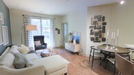 This single-floor residence offers a thoughtfully designed open-concept layout that seamlessly connects the living and dining areas into one cohesive, inviting space. Warm hardwood flooring runs throughout, grounding the interior with a sense of natural elegance and continuity.

The living area is bright and airy, with large sliding glass doors dressed in patterned curtains that allow generous amounts of natural light to fill the room while offering views of the surrounding exterior. A comfortable seating arrangement, anchored by a plush sofa and a classic leather accent chair, creates an ideal space for relaxation and conversation.

Adjacent to the living area, a compact dining nook provides an intimate setting for meals, featuring a round table and stylish chairs. A light wood credenza serves as both a functional storage piece and a display surface, accented by carefully chosen decorative objects that add character to the space.

A striking mixed-media wall art installation adds a bold, modern focal point to one wall, lending an artistic dimension to the overall aesthetic. Neutral, soft tones on the walls complement the warm flooring and furnishings, creating a balanced and welcoming atmosphere throughout. The layout is efficient yet spacious, making excellent use of every area within this single-level home.