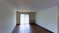 This single-floor space offers a clean and functional layout with a straightforward design. The main living area is defined by rich, dark hardwood flooring that extends throughout, providing a warm contrast to the light-toned walls and textured ceiling above. A prominent sliding glass door serves as a central architectural feature of the room, flanked by vertical blinds that allow for adjustable light control. The door floods the interior with generous natural light, creating a bright and airy atmosphere while also offering direct access to the outdoors. The neutral wall color and simple aesthetic keep the space feeling open and versatile, allowing for a variety of furniture arrangements and décor styles. Dark baseboards provide a subtle but defined border between the walls and flooring, adding a touch of visual structure to the room. The textured ceiling adds a layer of depth to the overall interior design. The space presents a practical and welcoming environment, well-suited for everyday living with its emphasis on natural light and a clean, unfussy aesthetic.