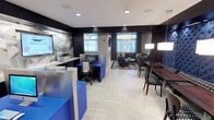 This well-appointed single-floor amenity space blends contemporary design with practical functionality, creating a welcoming environment suited for both work and relaxation. The open layout is anchored by light-toned porcelain tile flooring that flows seamlessly throughout, contrasted by striking dark charcoal and navy accents across cabinetry, walls, and furnishings.

A dedicated business center features vibrant royal blue workstations equipped with computers and a printer, complemented by an abstract graphic wall accent and ample task lighting. Adjacent to it, a dining area offers a rich wood communal table paired with patterned chairs and a dramatic tufted navy banquette wall, illuminated by pendant lighting that adds a sophisticated touch.

A refreshment station with a coffee maker and counter service is conveniently positioned near the main entrance, which features a glass-paneled door. The lounge area provides a more intimate retreat, centered around a linear fireplace set into a textured stone accent wall with a wall-mounted television above. A large window allows natural light to filter in, while a plush sectional sofa, a round coffee table, and a colorful accent chair complete the comfortable seating arrangement. Subtle damask-patterned wallcovering adds refined detail to the lounge walls, rounding out the space's polished, upscale aesthetic.