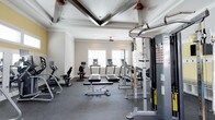 Located at 1680 Bluewater Way in Charleston, South Carolina, this well-appointed fitness center offers a comprehensive workout environment designed for both cardio and strength training. The space is defined by its striking architectural ceiling detail — a coffered tray design with elegant V-shaped wood beam accents and a beadboard inlay — giving the room a polished, upscale feel uncommon in typical gym settings. A ceiling fan centered within the tray keeps the space comfortable during workouts. Natural light flows in through multiple windows along the exterior wall, illuminating the room and creating an open, energizing atmosphere. The flooring consists of durable rubber gym matting, providing traction and cushioning throughout. Equipment includes multiple treadmills positioned beneath the windows, an elliptical trainer, a recumbent bike, a weight bench with free weights, and a multi-station cable and weight machine. The neutral color palette of soft whites and grays contributes to a clean, focused environment. Overall, this fitness center combines functional workout amenities with thoughtful architectural design, making it a standout community amenity in the Charleston area.