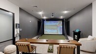 Located at 4126 River Rd in Wilmington, NC, this purpose-built indoor golf simulator suite offers a refined and functional environment for golf enthusiasts. The single-floor space is thoughtfully divided into two distinct zones that work seamlessly together. The simulator bay is the centerpiece of the suite, featuring a Full Swing Golf system complete with a ceiling-mounted projector, a large impact screen, and a high-quality artificial turf hitting mat. Protective netting lines the sides of the bay, and a golf club rack keeps equipment neatly organized. The dark accent walls in this area create an immersive, focused atmosphere ideal for practice and play. Adjacent to the simulator bay is a comfortable lounge and collaborative area finished with neutral-toned carpet and crisp white walls accented by a charcoal feature wall. This zone includes a sturdy wooden high-top table with leather bar stools, a sleek lounge chair paired with natural wood side tables, and a mounted display panel. A control kiosk provides easy access to the simulator's software. Black-framed glass doors and partitions add a modern, sophisticated touch, while recessed lighting and flush-mount fixtures ensure the entire space is well-lit. The overall design balances athletic utility with contemporary style.