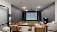 Located at 4126 River Rd in Wilmington, NC, this purpose-built indoor golf simulator suite offers a refined and functional environment for golf enthusiasts. The single-floor space is thoughtfully divided into two distinct zones that work seamlessly together. The simulator bay is the centerpiece of the suite, featuring a Full Swing Golf system complete with a ceiling-mounted projector, a large impact screen, and a high-quality artificial turf hitting mat. Protective netting lines the sides of the bay, and a golf club rack keeps equipment neatly organized. The dark accent walls in this area create an immersive, focused atmosphere ideal for practice and play. Adjacent to the simulator bay is a comfortable lounge and collaborative area finished with neutral-toned carpet and crisp white walls accented by a charcoal feature wall. This zone includes a sturdy wooden high-top table with leather bar stools, a sleek lounge chair paired with natural wood side tables, and a mounted display panel. A control kiosk provides easy access to the simulator's software. Black-framed glass doors and partitions add a modern, sophisticated touch, while recessed lighting and flush-mount fixtures ensure the entire space is well-lit. The overall design balances athletic utility with contemporary style.