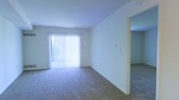 This single-floor space presents a straightforward and functional layout designed for comfortable everyday living. The main living area is finished with wall-to-wall carpeting in a neutral tone, providing a soft and cohesive foundation throughout the room. A sliding glass door fitted with vertical blinds serves as the focal point of the space, allowing an abundance of natural light to filter in while also providing direct outdoor access. The bright white walls contribute to an airy, open atmosphere, keeping the interior feeling fresh and uncluttered. A doorway visible along one wall leads to an adjoining room, suggesting a connected and practical floor plan. The smooth, white ceiling complements the clean lines of the walls, maintaining a consistent and minimalist aesthetic throughout. Overall, the space offers a simple, move-in-ready environment with neutral finishes that provide a versatile backdrop adaptable to a variety of interior styles and furnishing preferences.