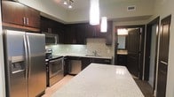This single-floor residence in the United States offers a thoughtfully designed interior with a strong emphasis on modern aesthetics and functional living. The kitchen serves as a central highlight, showcasing rich dark cabinetry that contrasts beautifully with light granite countertops. A large kitchen island provides generous prep and gathering space, anchored by two sleek cylindrical pendant lights that cast a warm, focused glow over the surface. The full suite of stainless steel appliances — including a refrigerator, range, microwave, and dishwasher — lends a polished, professional feel to the cooking space. A mosaic tile backsplash in neutral tones adds texture and visual interest between the countertops and upper cabinetry, while recessed track lighting illuminates the space from above. The kitchen sink is positioned along the perimeter cabinetry, maximizing workflow efficiency. A glimpse into an adjacent area suggests a bathroom with matching dark cabinetry and a well-appointed vanity, maintaining design continuity throughout. The overall ambiance of the space is contemporary and refined, with a cohesive color palette of dark wood tones, soft neutrals, and metallic finishes that together create an inviting yet sophisticated atmosphere.