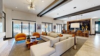 This well-appointed clubhouse spans a single floor of substantial square footage, offering a diverse range of amenity spaces designed for both socializing and productivity. The main lounge area serves as the heart of the clubhouse, featuring soaring ceilings with striking dark exposed beams, a contemporary chandelier, and expansive floor-to-ceiling sliding glass doors that flood the space with natural light and provide direct access to an outdoor patio. Plush sectional sofas, warm accent chairs, and a curated mix of patterned carpeting and polished stone flooring create an inviting, upscale atmosphere. Adjacent to the lounge is a sophisticated bar area framed by rich wood paneling, dramatic ring pendant lighting, and a large granite countertop with seating — complemented by a stone tile backsplash and built-in cabinetry. A dedicated game room offers a pool table, shuffleboard, dual wall-mounted TVs, and a warm wood accent wall with backlit shelving. A cozy lounge nook features built-in banquette seating beneath a striking stone-inspired wall mural, separated by a sleek barn door. The business center provides multiple workstations with computers and printing capabilities, while a private conference room includes a round wooden table, leather seating, a large display screen, and refined artwork throughout.