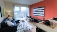 This contemporary single-floor residence offers a thoughtfully designed open-concept layout that seamlessly connects the living and dining areas. The living room is a standout space, anchored by a bold terracotta-orange accent wall adorned with a large-scale abstract artwork, creating a warm and vibrant focal point. Light hardwood-style flooring runs throughout, complementing the modern aesthetic and lending a sense of warmth and continuity across the space. A patterned area rug defines the seating area, which features a dark sectional sofa, a round coffee table, and an accent chair, all arranged to encourage conversation and relaxation. Floor-to-ceiling sliding glass doors flood the interior with abundant natural light and open directly onto a private balcony, offering elevated views of the surrounding urban landscape and treetops beyond. The dining area sits adjacent to the living space, reinforcing the open-plan design and making the layout ideal for both everyday living and entertaining. The overall ambiance is modern and stylish, with a curated mix of bold color, clean lines, and contemporary furnishings that give the space a vibrant yet inviting character.