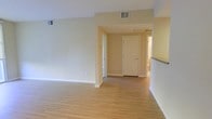 This single-floor residential space offers a bright and functional layout designed for comfortable everyday living. The main living area is finished with light hardwood-style flooring that extends throughout, creating a warm and cohesive aesthetic. Neutral, cream-toned walls complement the flooring and contribute to an airy, open atmosphere that feels welcoming and versatile.

The open floor plan allows for smooth transitions between the living space and adjacent areas, including what appears to be a kitchen partially visible to one side, featuring a granite or stone countertop edge. A central hallway connects the main living area to additional rooms, including what appears to be a bathroom visible at the end of the corridor. White panel interior doors provide clean, classic detailing throughout.

Ceiling-mounted HVAC vents are integrated discreetly into the layout, ensuring climate control without disrupting the clean visual lines of the space. Recessed wall outlets and neutral baseboards add to the tidy, well-maintained finish. The overall design emphasizes simplicity and practicality, with a light color palette and consistent materials that make the space feel open, uncluttered, and move-in ready.