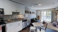 This single-floor space offers a thoughtfully designed open-concept layout that integrates the kitchen, dining, and living areas into one cohesive environment. The kitchen is outfitted with clean white cabinetry, brushed nickel hardware, a dark countertop, a stainless steel sink, and a stainless steel dishwasher, creating a modern and functional workspace. Track lighting overhead ensures the kitchen remains well-lit and practical. A floating shelf above adds a touch of personality with decorative accents. The dining area features a round marble-top table paired with classic black bentwood chairs and white stools, striking an elegant yet casual balance. Adjacent to the dining space, the living area provides a relaxed setting anchored by a comfortable sofa with plush cushions. A large window at the far end of the space floods the interior with natural light and offers a view of the outdoors, enhancing the sense of openness. Dark hardwood flooring flows throughout, grounding the light-toned walls and cabinetry with warmth and contrast. A staircase visible in the background hints at additional vertical access, while the overall layout maximizes every square foot with an efficient, welcoming design.