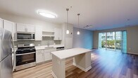 This well-appointed single-floor residence offers a thoughtfully designed open-concept layout that seamlessly connects the kitchen, dining, and living areas. The kitchen is a standout feature, equipped with stainless steel appliances including a gas range, microwave, and refrigerator, complemented by crisp white cabinetry, a grey tile backsplash, and a clean white countertop island with built-in shelving. Pendant lighting and a flush-mount ceiling fixture provide both task and ambient illumination throughout the space. The living area is bathed in natural light from floor-to-ceiling sliding glass doors that open to a private balcony, offering pleasant outdoor views. Hardwood-style flooring runs throughout the main living areas, while plush carpeting adds warmth to the bedroom, which features ample closet space with built-in shelving. The bathroom is finished with a white vanity, quartz-style countertop, undermount sink, chrome fixtures, and a large mirror with overhead lighting. A hallway with multiple doors connects the living areas to private rooms, offering a practical and efficient flow. The neutral color palette and modern finishes throughout create a clean, contemporary ambiance.