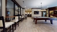 Located at 4000 City Walk Way in Charlottesville, Virginia, this single-floor community clubhouse offers a well-appointed gathering space designed with a cohesive, contemporary aesthetic. Bold dark wood accents, warm neutral tones, and striking yellow highlights carry throughout, creating a polished and inviting atmosphere.

The fully equipped kitchen and bar area serves as a central hub, featuring vibrant yellow countertops, dark cabinetry, stainless steel appliances, and stylish pendant lighting. Bar-height seating along the island makes it ideal for casual socializing or community events.

A comfortable lounge area offers plush seating arranged around a low coffee table, bathed in natural light from large floor-to-ceiling windows that look out onto the surrounding building exterior. Soft area rugs and layered lighting add warmth to this relaxing retreat.

A dedicated nook with a modern fireplace framed in yellow mosaic tile provides an intimate seating area, complemented by floating shelving, sculptural décor, and a striped upholstered banquette on the adjacent wall.

The game and entertainment area features a full-size billiards table beneath an angular pendant light, a wall-mounted flat-screen TV, and high-top bar seating. A separate business center with individual workstations is conveniently accessible from this room, rounding out the versatile amenities this clubhouse has to offer.