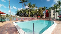 This outdoor pool area offers a inviting tropical retreat, designed for relaxation and leisure. The centerpiece is a generously sized swimming pool with clear, turquoise water, accessible via a chrome handrail entry. The pool is surrounded by a well-maintained deck finished in warm terracotta-toned pavers, providing ample space for movement and lounging.

A row of red-framed lounge chairs lines the far edge of the pool deck, offering dedicated sunbathing areas for guests or residents. A white metal perimeter fence encloses the space, ensuring safety while maintaining an open, airy feel. Red market umbrellas are positioned toward the back of the deck, providing shaded seating options.

The surrounding landscape is defined by tall, mature palm trees and dense tropical foliage, creating a lush, resort-like backdrop. The natural canopy contributes both aesthetic appeal and partial shade to the area. A multi-story building is partially visible in the background, indicating this pool serves a larger residential or hospitality complex. The overall ambiance is bright, relaxed, and unmistakably tropical, ideal for warm-weather enjoyment under open blue skies.