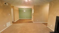 This single-floor space offers a welcoming and functional layout designed for comfortable everyday living. The open-concept living and dining area serves as the heart of the space, seamlessly blending two distinct zones through a thoughtful transition from plush wall-to-wall carpeting in the living area to smooth wood-look hard flooring in the dining area. The warm beige walls provide a neutral, inviting backdrop throughout, while a sage green accent wall in the dining area adds a subtle pop of color and visual depth. Lighting is a notable feature, with an elegant pendant light fixture centered over the dining area and a chandelier gracing the living space, both contributing to a warm and ambient atmosphere. Clean white baseboards provide a crisp contrast against the flooring and walls, lending a polished finish to the interior. A glimpse of an adjacent area — likely a kitchen or utility space — is visible through an opening, suggesting a practical and connected floor plan. The overall ambiance is clean, neutral, and move-in ready, offering a versatile canvas well-suited to a variety of interior styles and furnishing choices.