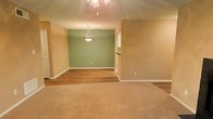 This single-floor space offers a welcoming and functional layout designed for comfortable everyday living. The open-concept living and dining area serves as the heart of the space, seamlessly blending two distinct zones through a thoughtful transition from plush wall-to-wall carpeting in the living area to smooth wood-look hard flooring in the dining area. The warm beige walls provide a neutral, inviting backdrop throughout, while a sage green accent wall in the dining area adds a subtle pop of color and visual depth. Lighting is a notable feature, with an elegant pendant light fixture centered over the dining area and a chandelier gracing the living space, both contributing to a warm and ambient atmosphere. Clean white baseboards provide a crisp contrast against the flooring and walls, lending a polished finish to the interior. A glimpse of an adjacent area — likely a kitchen or utility space — is visible through an opening, suggesting a practical and connected floor plan. The overall ambiance is clean, neutral, and move-in ready, offering a versatile canvas well-suited to a variety of interior styles and furnishing choices.
