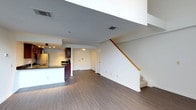 This two-floor space offers a thoughtfully designed layout that balances open living with functional design. The main level features an open-concept floor plan where the living area flows seamlessly into the kitchen. The kitchen is equipped with dark-toned cabinetry, a stainless steel refrigerator, a dark countertop peninsula, and track lighting that provides focused illumination over the workspace. The flooring throughout the main level is a warm-toned hardwood-style laminate that adds a sense of continuity and warmth to the space. Crisp white walls and clean lines give the interior a bright, modern aesthetic. A staircase with a natural wood handrail connects the main level to the upper floor, adding a classic architectural touch. Overhead, HVAC vents are integrated into the ceiling, ensuring year-round comfort. The open layout of the main level creates a generous sense of space, making it well-suited for both everyday living and entertaining. The second floor provides additional living or sleeping quarters, expanding the overall functionality of the home. The combination of practical finishes, clean design, and a well-connected two-floor layout makes this a versatile and comfortable space.