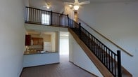 This two-story residence offers a thoughtfully designed open-concept layout that maximizes both space and functionality. The main level centers around a generous living area finished with wood-look flooring and neutral walls that create a bright, welcoming atmosphere. A kitchen sits adjacent to the living space, featuring warm wood cabinetry, a white refrigerator, a sink with a countertop overhang, and track lighting — making it both practical and accessible. A sliding glass door at the rear of the main level allows natural light to filter into the living area and provides access to the outdoors. A staircase with a light wood handrail and sleek black metal balusters leads to the upper loft level, which overlooks the main living area below. The loft features the same black metal railing system, reinforcing a cohesive design aesthetic throughout the home. Ceiling fans on both levels provide year-round comfort and contribute to the home's casual yet polished character. The open vertical space created by the two-story design gives the interior an expansive, airy quality, making this a distinctive and comfortable multi-level home.