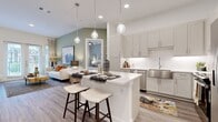 This single-floor residence offers a thoughtfully designed open-concept layout accommodating two bedrooms and two full bathrooms. The combined kitchen and living area forms the heart of the home, where light wood-tone flooring, vaulted ceilings, and large glass French doors flood the space with natural light and provide outdoor views. The kitchen is outfitted with gray shaker-style cabinetry, white quartz countertops, a white penny-tile backsplash, stainless steel appliances including a French door refrigerator, and a central island with seating for two, illuminated by globe pendant lights. Built-in open shelving flanks a decorative accent wall, adding functional display space. The living area is generously proportioned, offering room for comfortable seating arrangements. The primary bedroom is a well-sized retreat with direct access to a private en-suite bathroom featuring a dual-sink vanity with white quartz countertops, a glass-enclosed shower, and a walk-in closet with built-in shelving. The second bedroom receives natural light through a large window and is served by a second full bathroom with a combination tub and shower. Wood-look flooring runs consistently throughout the entire residence, contributing to a cohesive, contemporary aesthetic.