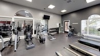 This fitness center offers a comprehensive workout environment thoughtfully organized across a single floor. The space is finished in a contemporary style, with neutral gray walls, durable rubber flooring, and recessed panel lighting that ensures consistent, bright illumination throughout. Large windows along one wall allow natural light to pour in and provide views of surrounding greenery, creating an open and inviting atmosphere during workouts.

The cardio section is well-appointed with multiple treadmills, elliptical trainers, a stair climber, and a recumbent bike, giving users a variety of options for aerobic exercise. An arched window accent near the cardio area adds an architectural detail that softens the utilitarian nature of the space. The strength-training area includes a multi-station cable machine, a dedicated free-weight zone with a full rack of dumbbells in graduated sizes, and adjustable benches for targeted resistance work.

Wall-mounted mirrors are strategically placed to visually expand the space and allow users to monitor their form. A wall-mounted television provides entertainment. A water fountain station is conveniently accessible, and posted fitness guides offer workout reference information. The facility connects to an adjacent room, extending its overall usability.