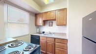 This single-floor unit is thoughtfully laid out, offering a comfortable and functional living environment. The main living area is finished with plush wall-to-wall carpeting and neutral-toned walls, with a large window fitted with vertical blinds that allows natural light to flow in while providing outdoor views of surrounding greenery. A distinct blue accent wall adds a subtle visual element to the space. The layout connects the living area to the kitchen and private rooms via a central hallway, providing clear separation between shared and private spaces.

The kitchen is equipped with warm honey-toned wood cabinetry extending from base to upper wall level, offering generous storage capacity. Dark black appliances, including a coil-top range and dishwasher, contrast with the light laminate countertops. A single-basin sink is positioned beneath upper cabinets, and a window brings in natural light. The flooring transitions to hardwood-style material in this area.

The bedrooms are carpeted and bright, each featuring windows with vertical blinds and ample wall space. Multiple closets and connecting doors provide practical storage and accessibility. The bathroom includes tile wall surround, a vanity with a white countertop, a dark wood cabinet base, a mirrored medicine cabinet, and a shower-tub combination. Overall, the unit presents a clean, move-in ready environment with consistent finishes throughout.