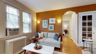 This single-floor residence offers a thoughtfully designed layout with distinct spaces that balance character and functionality. The living room serves as the focal point, featuring rich warm tones with a bold burnt-orange accent wall adorned with framed artwork and wall sconces that provide a soft, ambient glow. Large windows allow natural light to fill the room, complementing the warm hardwood flooring that runs throughout. A marble-topped coffee table and a neutral sofa anchor the space, while an elegant archway connects the living area to the rest of the unit.

The kitchen is well-appointed with rich cherry wood cabinetry, granite countertops, and a full suite of stainless steel appliances, including a gas range, built-in microwave, dishwasher, and refrigerator, offering ample storage and workspace.

Two bedrooms provide comfortable retreats, each featuring hardwood floors, neutral wall tones, and windows that overlook greenery outdoors. The primary bedroom offers generous space for a dresser and seating area, while the second bedroom functions equally well as a guest room or home office.

The bathroom retains a classic retro aesthetic, with warm yellow square tile accents, black trim detailing, white tile flooring, and a large wall mirror above a pedestal-style vanity, preserving a timeless vintage charm.