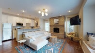 This well-appointed single-floor home offers an open-concept layout that seamlessly connects the living, dining, and kitchen areas, creating a spacious and inviting atmosphere throughout. Rich hardwood flooring runs continuously across the space, anchoring each area with warmth and cohesion.

The living room serves as the heart of the home, centered around a striking full-height brick fireplace that adds rustic character and charm. A plush white sectional sofa, vibrant Persian-style area rug, and a contemporary multi-globe gold chandelier combine to create an elegant yet comfortable gathering space. A built-in window seat with decorative pillows offers a cozy nook bathed in natural light from large windows.

The kitchen is thoughtfully designed with crisp white shaker cabinetry, a mosaic tile backsplash, stainless steel appliances, and a white peninsula with gold-accented bar stools — perfect for casual dining or entertaining. Elegant pendant lighting adds a refined touch overhead.

Adjacent to the living area, the dining space accommodates a full dining table and chairs, with a set of double doors that introduce additional natural light. The textured ceilings and recessed lighting throughout provide a clean, finished look, while multiple windows ensure the entire home remains bright and airy.