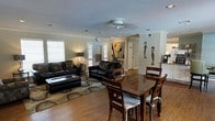 This single-floor space showcases an open-concept layout that seamlessly connects the living and dining areas, creating a spacious and functional environment. The flooring throughout is warm hardwood, adding a classic and inviting character to the space.

The living area is furnished with two dark leather sofas arranged around a rectangular coffee table, all set atop a patterned area rug. A ceiling fan with a light fixture is centrally positioned overhead, and crown molding lines the walls, lending a polished, finished appearance. Natural light filters in through two windows fitted with horizontal blinds on the left wall.

The dining area features a solid wood table accompanied by matching wooden chairs with upholstered seats. Adjacent to the dining space, a small counter or bar area is visible, suggesting a connection to the kitchen. Decorative elements throughout include a bold abstract painting, two sculptural African-style figurines, and a table lamp, giving the space a curated, artistic feel. The neutral wall tones provide a clean backdrop that complements the rich wood tones and dark furnishings throughout.