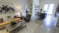This single-story space showcases an open-plan layout that seamlessly connects the living and dining areas, creating a cohesive and functional environment. The flooring transitions between polished concrete and light wood-toned surface treatments, lending the space an industrial yet warm character. The living area is anchored by a tan sofa and a distinctive wooden glider chair in deep green upholstery, complemented by a rustic coffee table displaying decorative accessories and greenery. A table lamp provides warm accent lighting, enhancing the inviting atmosphere. A freestanding open shelving unit divides the space visually while displaying books, plants, and decorative objects. The dining area features a dark bistro-style table with matching black chairs, positioned near a window that draws in natural light. Decorative metal numbers mounted on the wall serve as a notable design feature. A doorway visible in the background hints at a bedroom beyond, suggesting a practical flow throughout the unit. The overall aesthetic blends rustic, industrial, and eclectic influences, resulting in a characterful and comfortable interior.