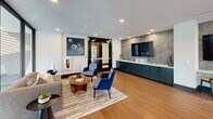 Located at 5580 Farrington Rd in Durham, NC, this single-floor amenity space offers a well-appointed environment divided between a social lounge and a dedicated fitness center. The lounge area makes a strong first impression with floor-to-ceiling black-framed windows that flood the interior with natural light and provide open views of the surrounding community. Rich wood-paneled accent columns anchor the space, complementing the wood-look tile flooring and neutral-toned furnishings, including a sectional sofa and accent chairs arranged around a round coffee table. A pool table adds a recreational focal point, while a built-in media console set against a textured mosaic tile wall provides an entertainment hub near the entrance. Contemporary artwork and recessed lighting complete the polished aesthetic. The fitness center is equally impressive, featuring large windows on multiple walls that bring in abundant daylight and outdoor views. Sleek light-wood flooring, large wall mirrors, and ceiling fans create an energizing workout environment. The space is outfitted with cycling bikes, rowing machines, treadmills, kettlebells, medicine balls, and stability equipment, catering to a wide range of fitness routines. A glass railing offers a visual connection between workout zones, enhancing the open, airy feel throughout.