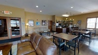 This single-floor space serves as a comfortable gathering area that seamlessly blends a casual lounge with a café-style refreshment station. The lounge area is anchored by a rich brown leather sofa and a cream-colored loveseat, arranged around a patterned area rug and a central coffee table, creating an inviting spot for relaxation and conversation. Warm yellow-toned walls contribute to the cozy, welcoming ambiance throughout the space.

Adjacent to the lounge, a dining area features multiple wooden tables paired with mixed seating, ideal for casual meals or social gatherings. The café station is well-appointed with a coffee machine, microwave, stainless steel refrigerator, and open shelving stocked with supplies — all illuminated by a row of pendant lights that add a warm, functional glow to the area. Recessed lighting across the ceiling ensures the entire space is evenly and brightly lit.

Decorative touches, including framed artwork and a coffee-themed sign on the walls, reinforce the casual and community-oriented character of the space. A dark hardwood-style floor runs throughout, tying together the lounge, dining, and café zones into a cohesive, functional environment well-suited for everyday use and community interaction.