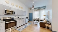 Located in Getzville, NY, this single-floor unit offers four bedrooms and four bathrooms within a efficiently designed layout. Light wood flooring runs throughout, complementing the clean, contemporary aesthetic of the space.

The open-concept kitchen and living area serves as the heart of the unit. The kitchen is outfitted with stainless steel appliances — including a refrigerator, electric range, microwave, and dishwasher — paired with granite countertops and sleek gray cabinetry. Tall ceilings with exposed ductwork lend an industrial character to the living area, which features large windows that invite ample natural light. A bold blue entry door adds a distinctive pop of color.

Each of the four bedrooms is a private, cozy space with its own dedicated bathroom — a standout feature of this unit. The bedrooms include open closets with wire shelving, providing practical storage solutions. The bathrooms are clean and functional, each equipped with a vanity featuring gray cabinetry, a granite countertop, and a walk-in shower with curtain.

A connecting hallway ties the rooms together in a logical flow. The combination of modern finishes, individual en-suite bathrooms, and a shared social space makes this unit well-suited for multi-occupant living.