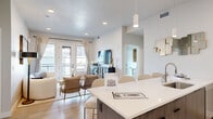 This thoughtfully designed single-floor residence showcases a seamless open-concept layout that connects the kitchen, dining, and living areas into one cohesive, light-filled space. The kitchen is a standout, featuring a large center island with a white quartz countertop, an undermount sink, pendant lighting, rich dark wood cabinetry, and a full suite of stainless steel appliances including a French door refrigerator, electric range with microwave, and dishwasher. Upper white cabinetry adds contrast and ample storage.

The living area is bright and welcoming, anchored by French doors that open to an outdoor balcony, inviting abundant natural light. Light wood-tone flooring runs throughout the main living spaces, complementing the neutral palette. A secondary sitting area offers a cozy retreat with large windows and elegant décor accents.

The bedroom is generously sized with plush carpeting, a large window framing outdoor views, and direct access to a spacious walk-in closet featuring built-in shelving and hanging rods. The bathroom is clean and modern, with a floating dark wood vanity, white quartz countertop, and an undermount sink. A dedicated laundry area with in-unit appliances adds everyday convenience. The overall ambiance is contemporary, sophisticated, and move-in ready.