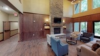 This single-floor communal space is thoughtfully designed to create a warm and inviting atmosphere for gathering and relaxation. The centerpiece of the main lounge area is a dramatic floor-to-ceiling stacked stone fireplace, complete with a wood mantel and a wall-mounted television above, drawing the eye upward toward the soaring vaulted ceiling painted in a soft golden yellow. Rich wood paneling lines the walls throughout, complemented by wood-look tile flooring that adds warmth and continuity across the space. Built-in shelving with subtle accent lighting provides decorative display areas, while large windows along one wall allow natural light to filter in and offer views to the exterior. Comfortable seating, including upholstered chairs and sofas, is arranged to encourage conversation around the fireplace. A kitchenette area is integrated into the space, featuring warm wood cabinetry mounted against a stacked stone accent wall, a stainless steel sink, countertop workspace, and under-counter refrigeration — ideal for hosting events or casual refreshments. A high-top table with bar stools provides an informal dining spot nearby. The lobby area includes elevator access and additional high-top seating, reinforcing the space's function as a well-appointed, accessible communal hub.